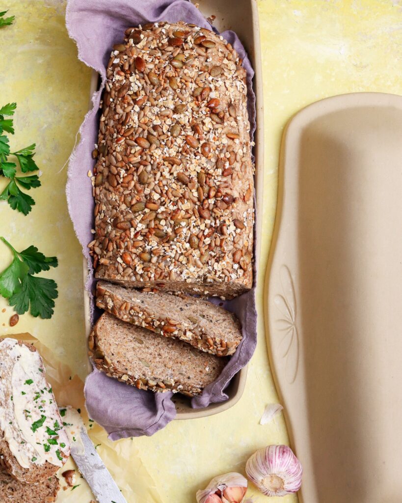 Fresh whole grain bread in artisan covered bread pan with garlic and spread on side.